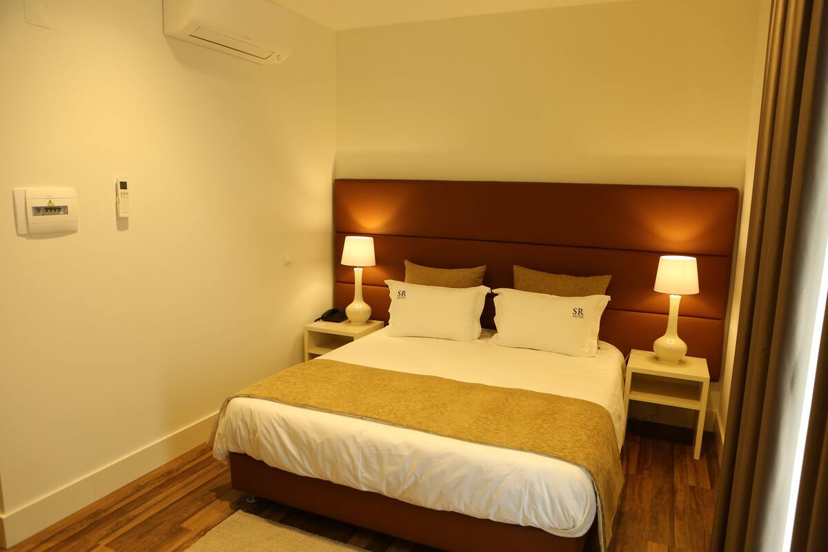 A hotel room with a large bed, two bedside tables with lamps, a wall-mounted air conditioner, and a power outlet.