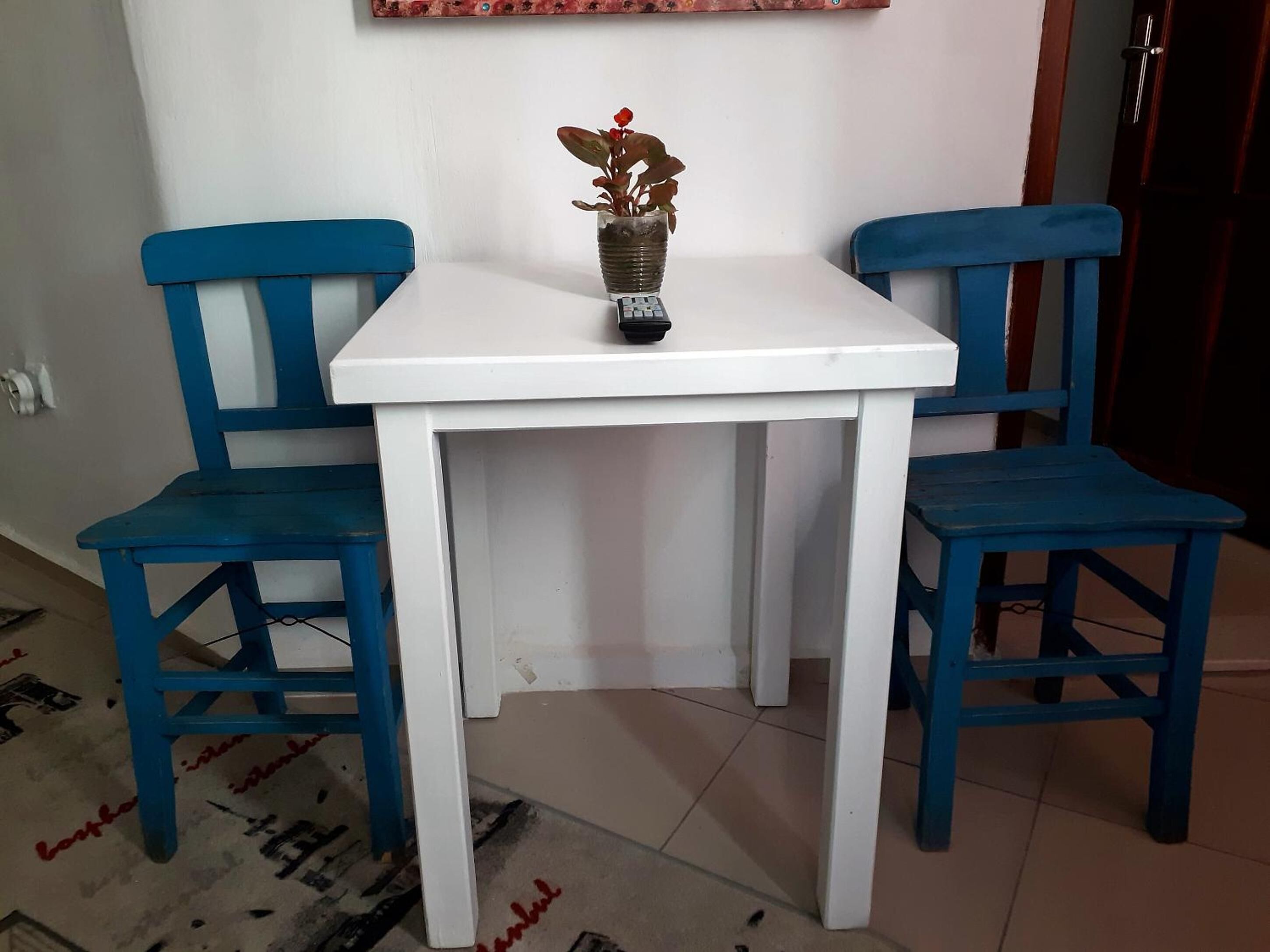 A small white table with two blue chairs, a potted plant, and a wall-mounted shelf with a decorative item.