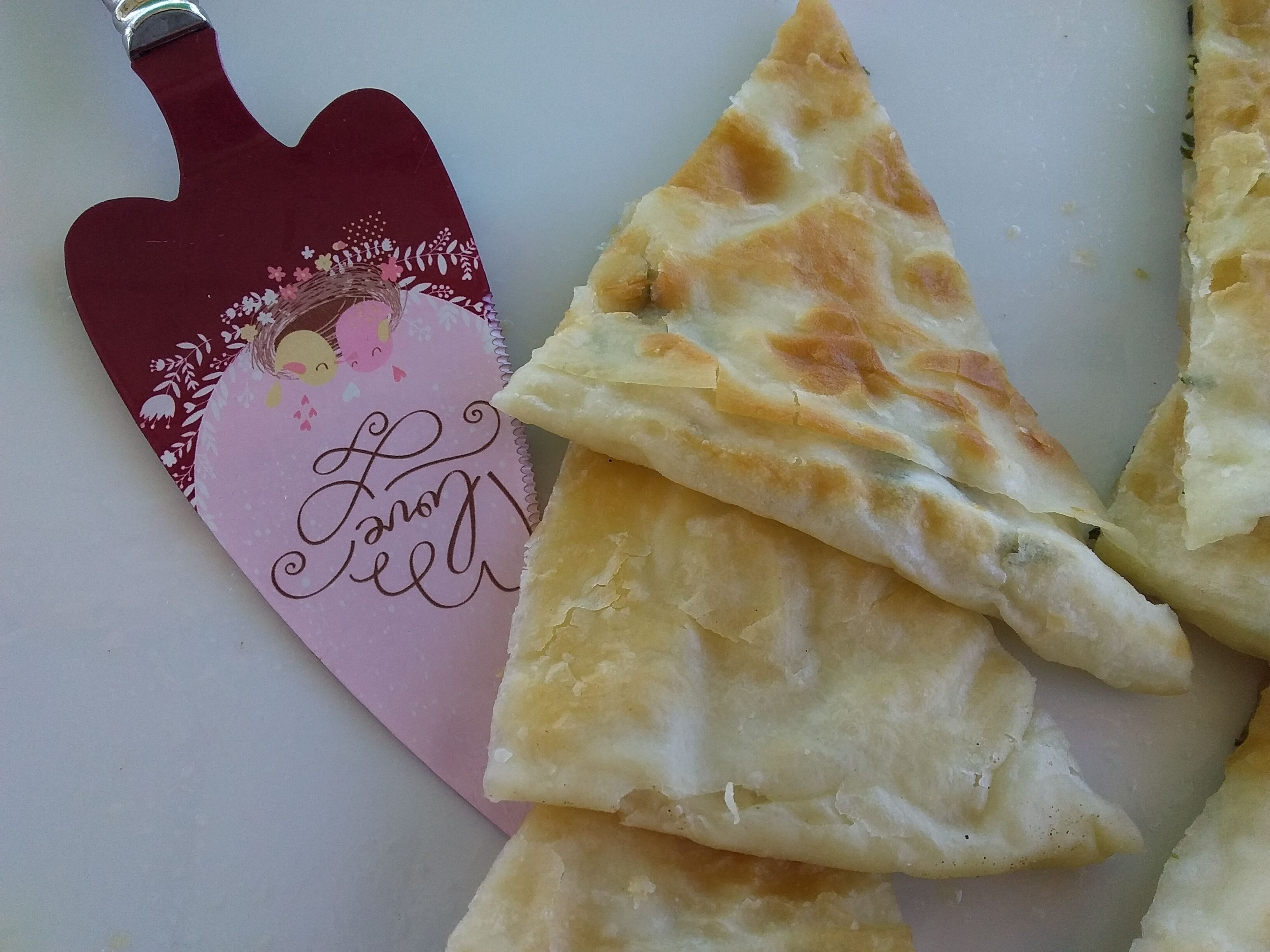 Un cortapizza con un mango decorativo y una porción de pizza sobre una superficie blanca.