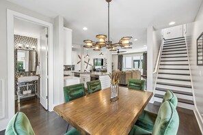 A modern kitchen with white cabinets and a central island, a dining area with a wooden table and green chairs, and a staircase with a railing.