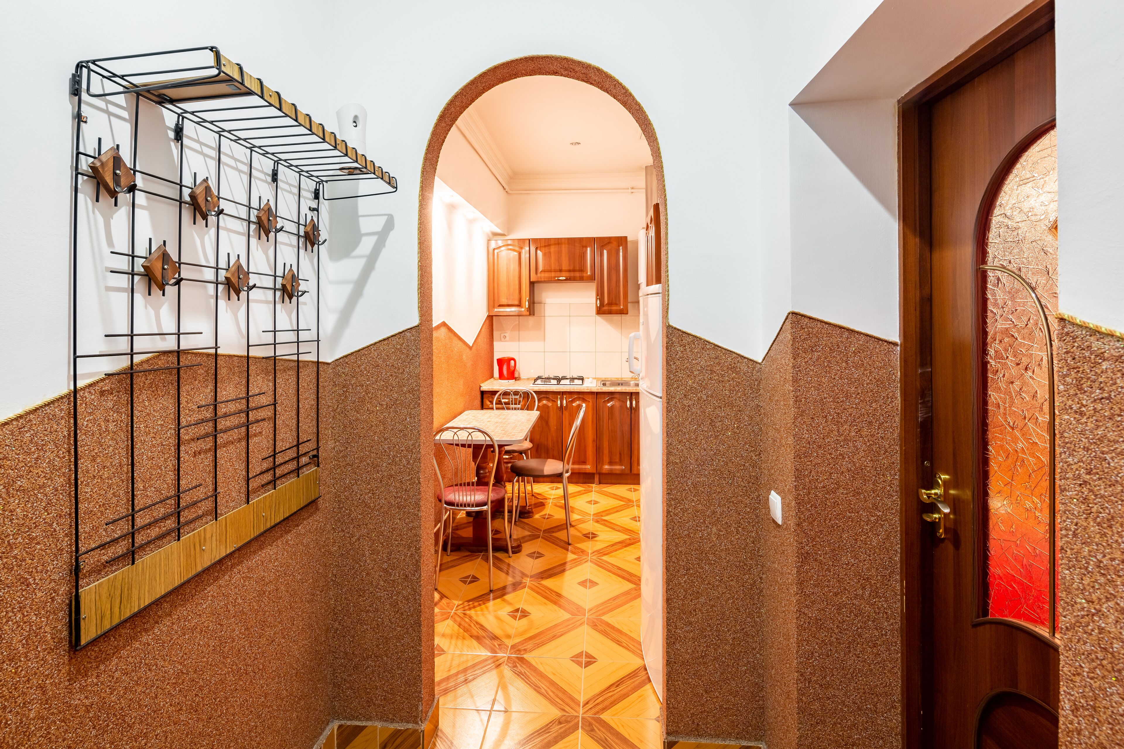 A narrow hallway with a tiled floor, a wooden door, and a kitchen visible through an archway.