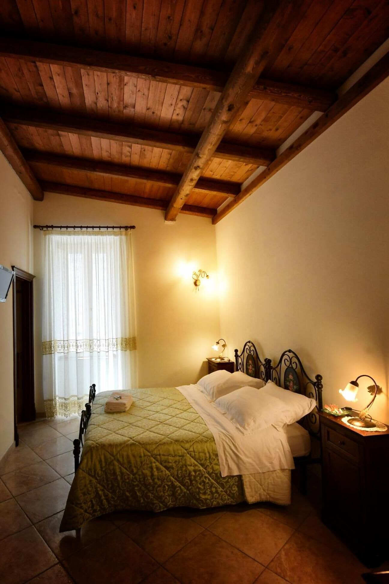 A bedroom with a wooden ceiling, a bed with a quilt, two bedside lamps, and a window with curtains.