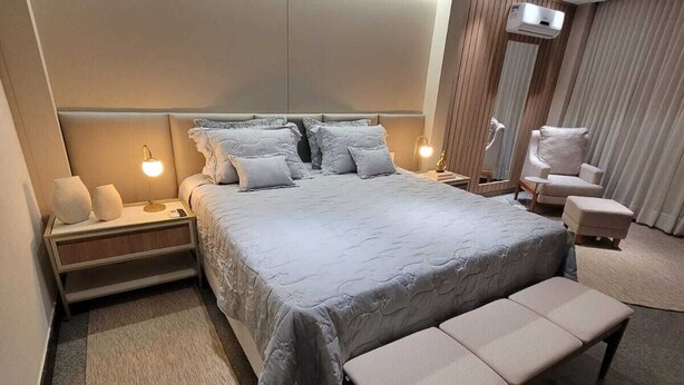A modern hotel bedroom with a large bed, bedside lamps, a bench, a chair, and a small table. - Lindoya Hotel (Catalao)