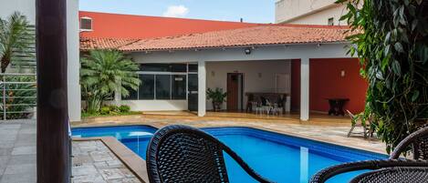 A swimming pool with wicker chairs, a covered patio, and a building with a red roof.