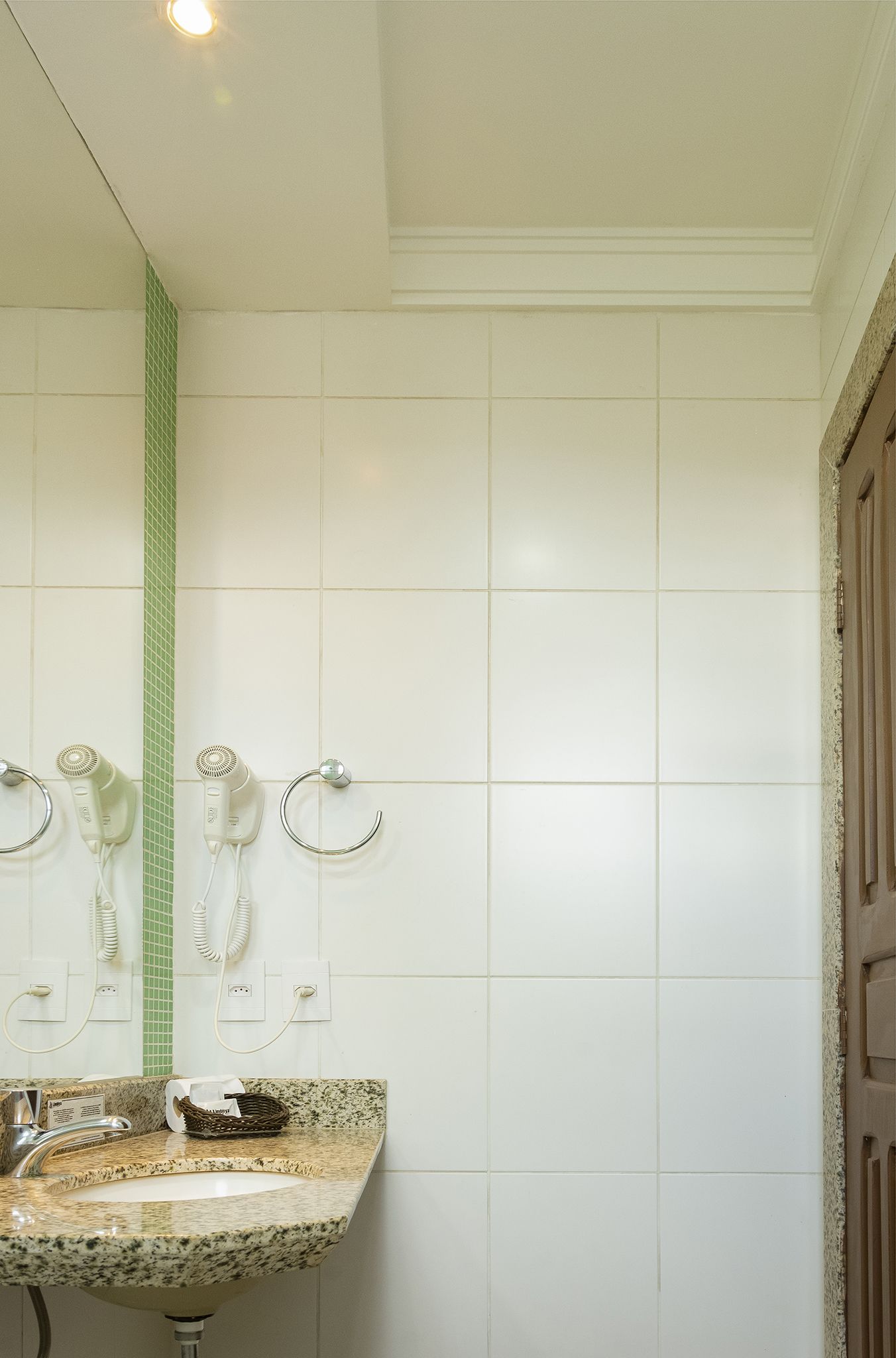 A bathroom with white tiled walls, a granite sink, and two hair dryers mounted on the wall.