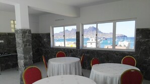 Een eetgedeelte met ronde tafels en rode stoelen, grote ramen met uitzicht op zee en bergen, en stenen muren.