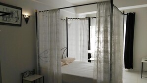 Een bed met hemelbed, met een witte matras en kussens, een klein nachtkastje en een stoel.