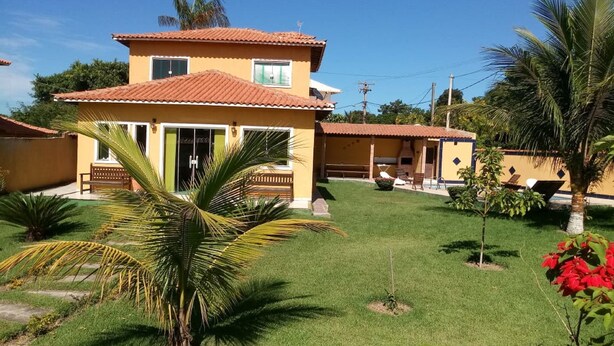 A two-story house with a tiled roof, a small patio with a bench, and a well-maintained lawn with palm trees and red flowers. - Casa em Armação de Búzios (Búzios)