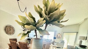 A living room with a potted plant, a clock, and a wooden tray with books. - July 27-Aug 1 open.  Modern Condo—Best location—beach, Ft Clinch & downtown. (Fernandina Beach)