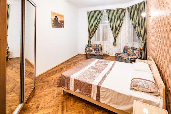 A bedroom with a bed, two chairs, a wooden floor, a mirror, and a picture on the wall.