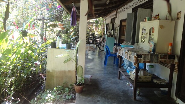 A covered outdoor area with a concrete floor, potted plants, a blue chair, and a refrigerator. - Grandma's Home Sangkhlaburi (Sangkhla Buri)