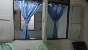 A room with a window, two green plastic chairs, a small table with a potted plant, and a wooden bench. - Grandma's Home Sangkhlaburi (Sangkhla Buri)