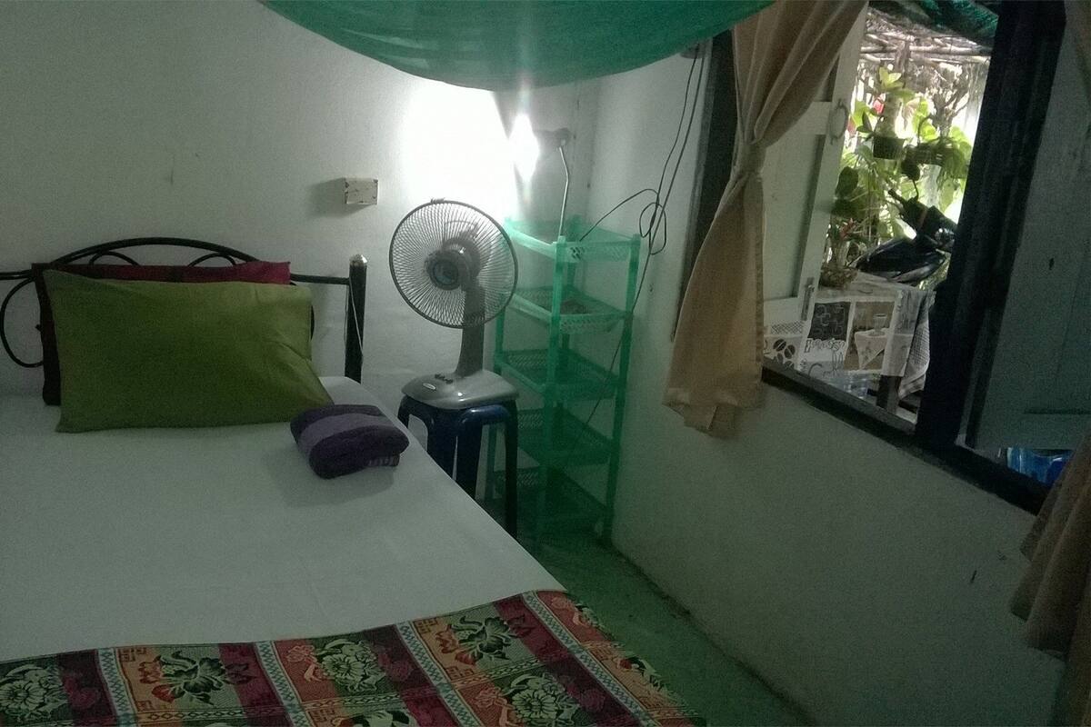 Um quarto pequeno e modesto, com uma cama, um ventilador e uma janela com cortinas.