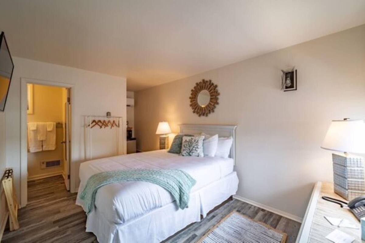 A bedroom with a bed, bedside lamps, a wall clock, and a door leading to a bathroom.