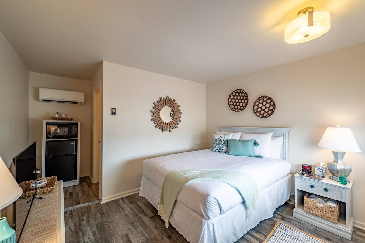 A bedroom with a bed, bedside table, lamp, wall decor, and a small kitchenette.
