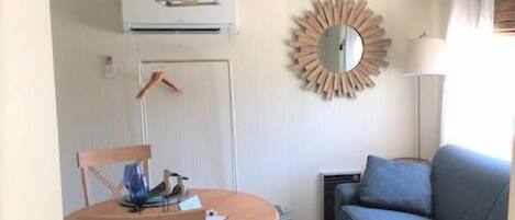 A compact living space with a round wooden dining table, two wooden chairs, a blue sofa, a round mirror, and a hanging light fixture.