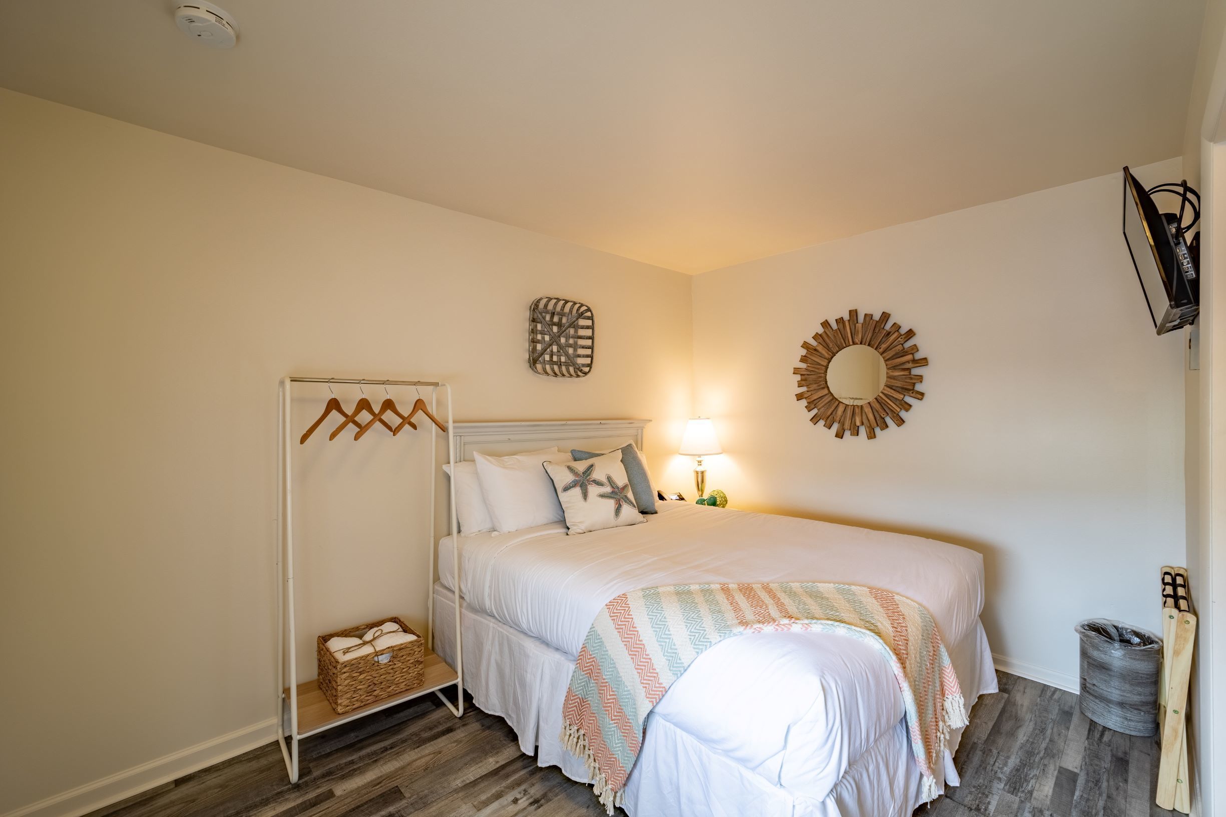 A bedroom with a bed, a bedside table, a lamp, a wall-mounted mirror, and a coat rack.