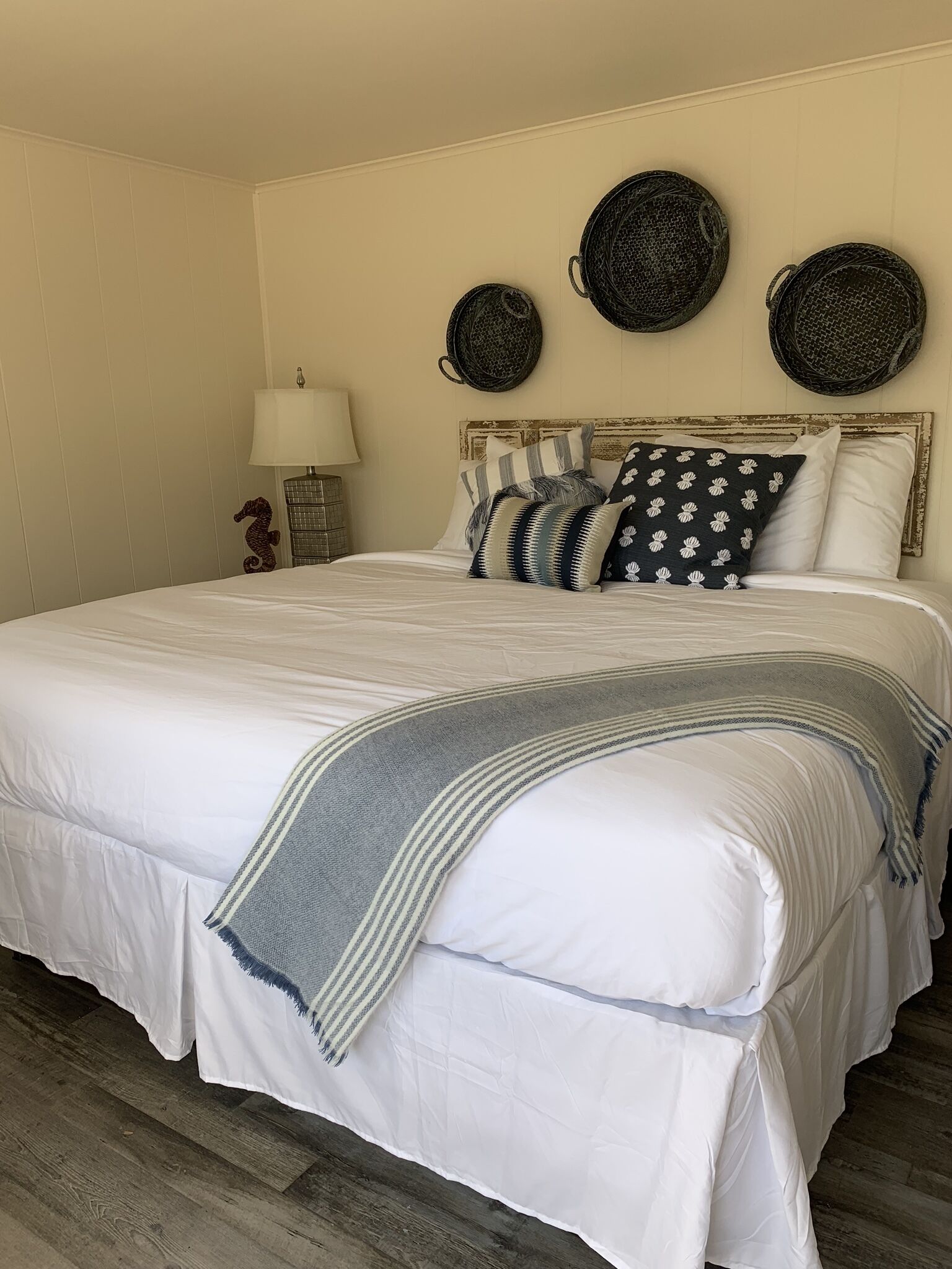 A neatly made bed with a grey throw blanket, a wooden nightstand with a lamp, and three woven baskets on the wall.