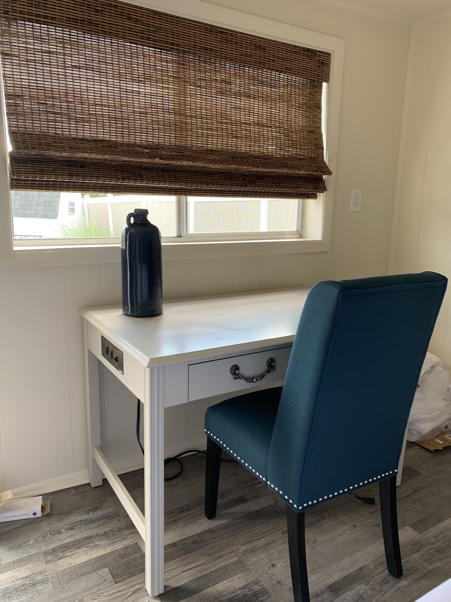 Un escritorio blanco con una silla azul, una botella de agua negra y una ventana con persiana marrón.