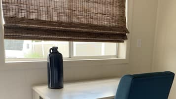Un escritorio blanco con una silla azul, una botella de agua negra y una ventana con persiana marrón.