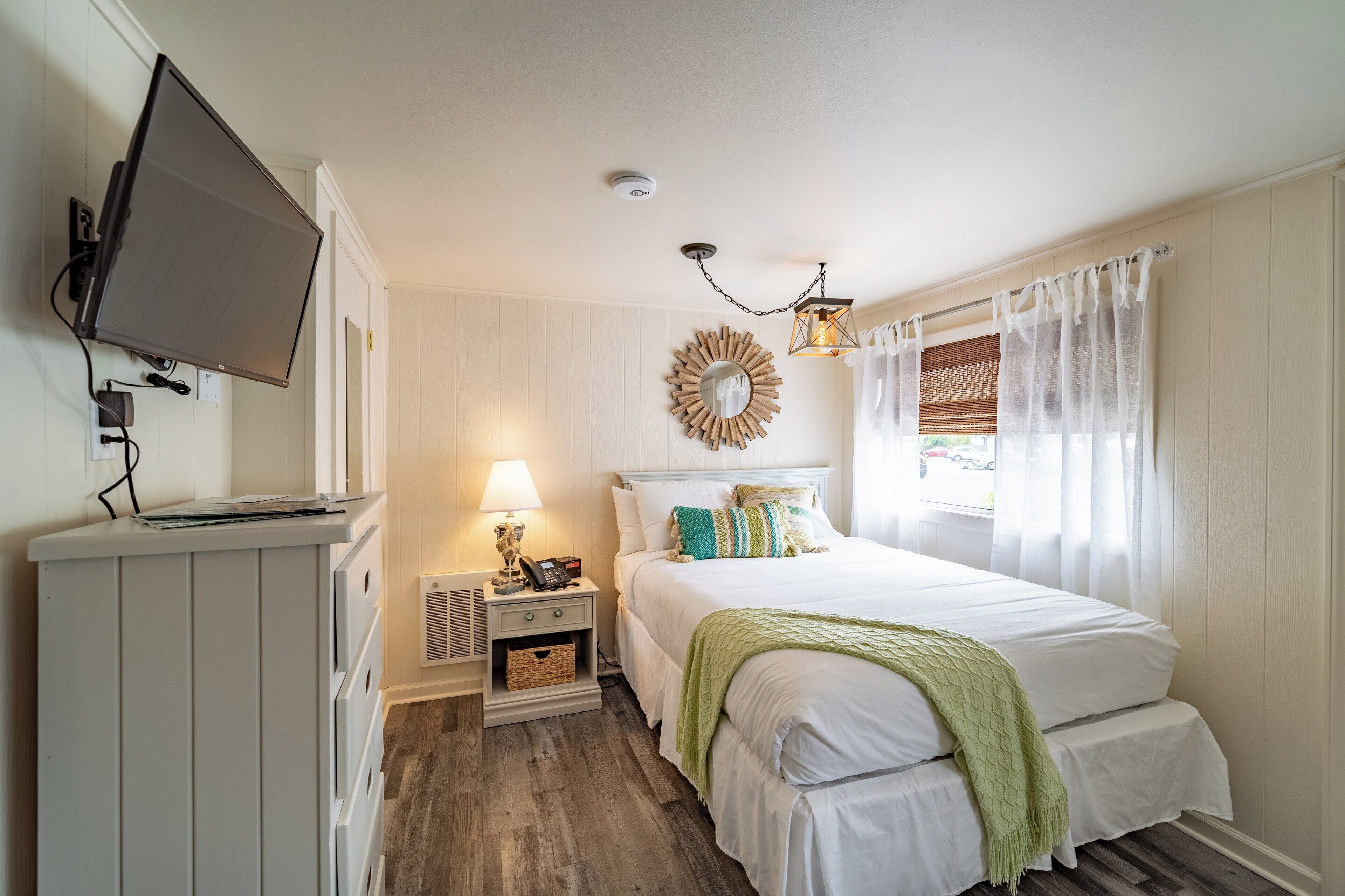 Quarto com cama, mesa de cabeceira com luminária, televisão na parede e janela com cortinas.
