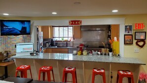 Una cocina con armarios de madera, encimera de granito y taburetes rojos. Hay un televisor montado en la pared, una ventana con persianas y una señalización que indica 'EXIT'.