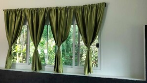 Habitación con ventana, cortinas verdes, cabecera oscura y vista a plantas afuera.