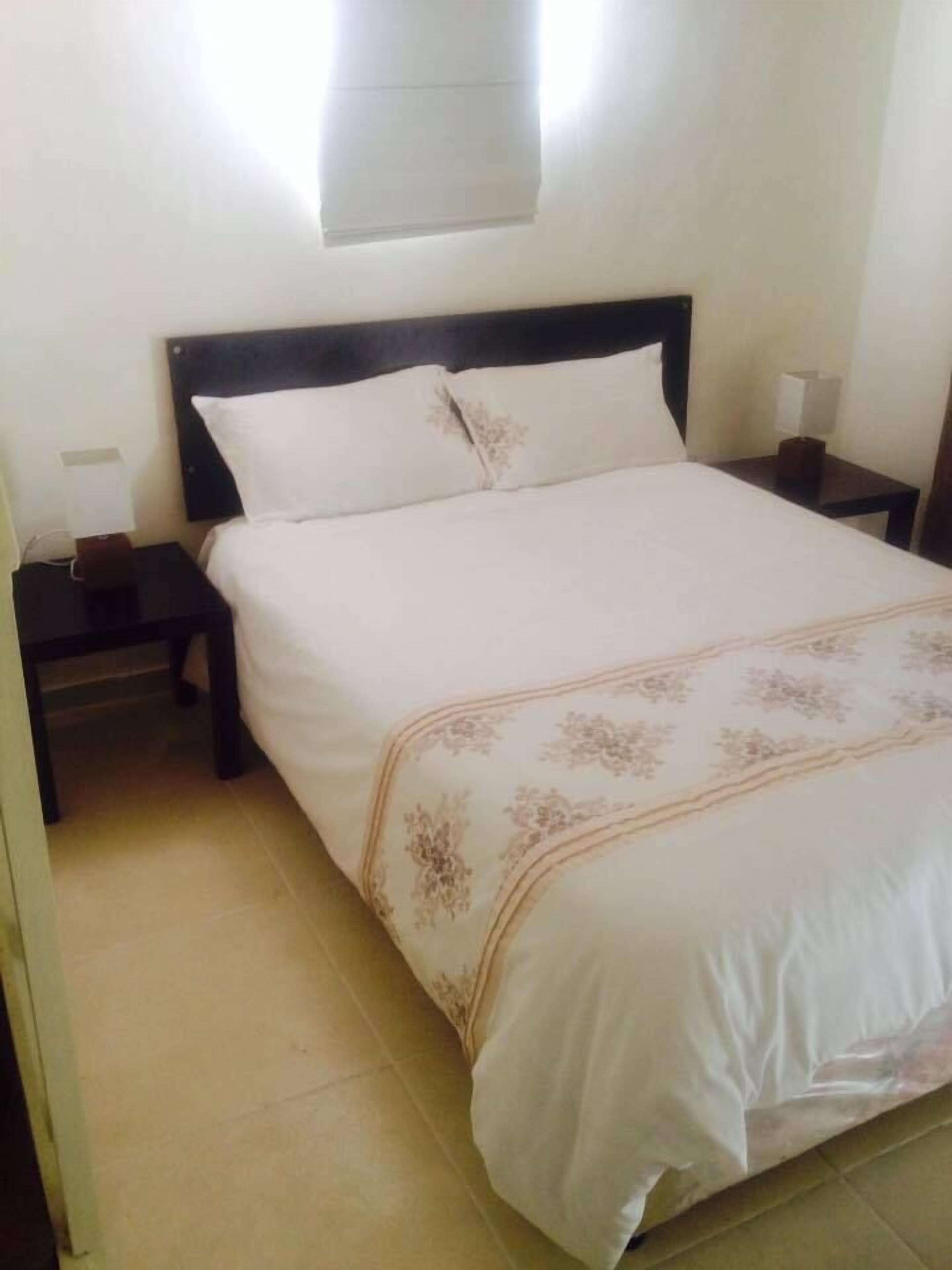 A neatly made bed with a floral-patterned bedspread, a bedside table with a lamp, and a wall-mounted light fixture.