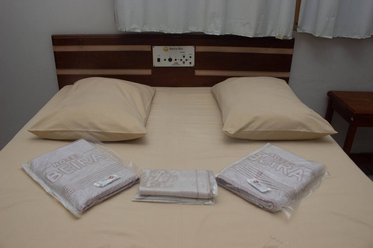 Ein Bett mit hölzernem Kopfteil, zwei beigefarbene Kissen und drei gefaltete Decken mit dem Aufdruck 'Hotel Beira'.