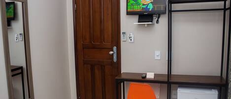 Una habitación con puerta de madera, televisor montado en la pared, escritorio con silla y un refrigerador pequeño blanco.