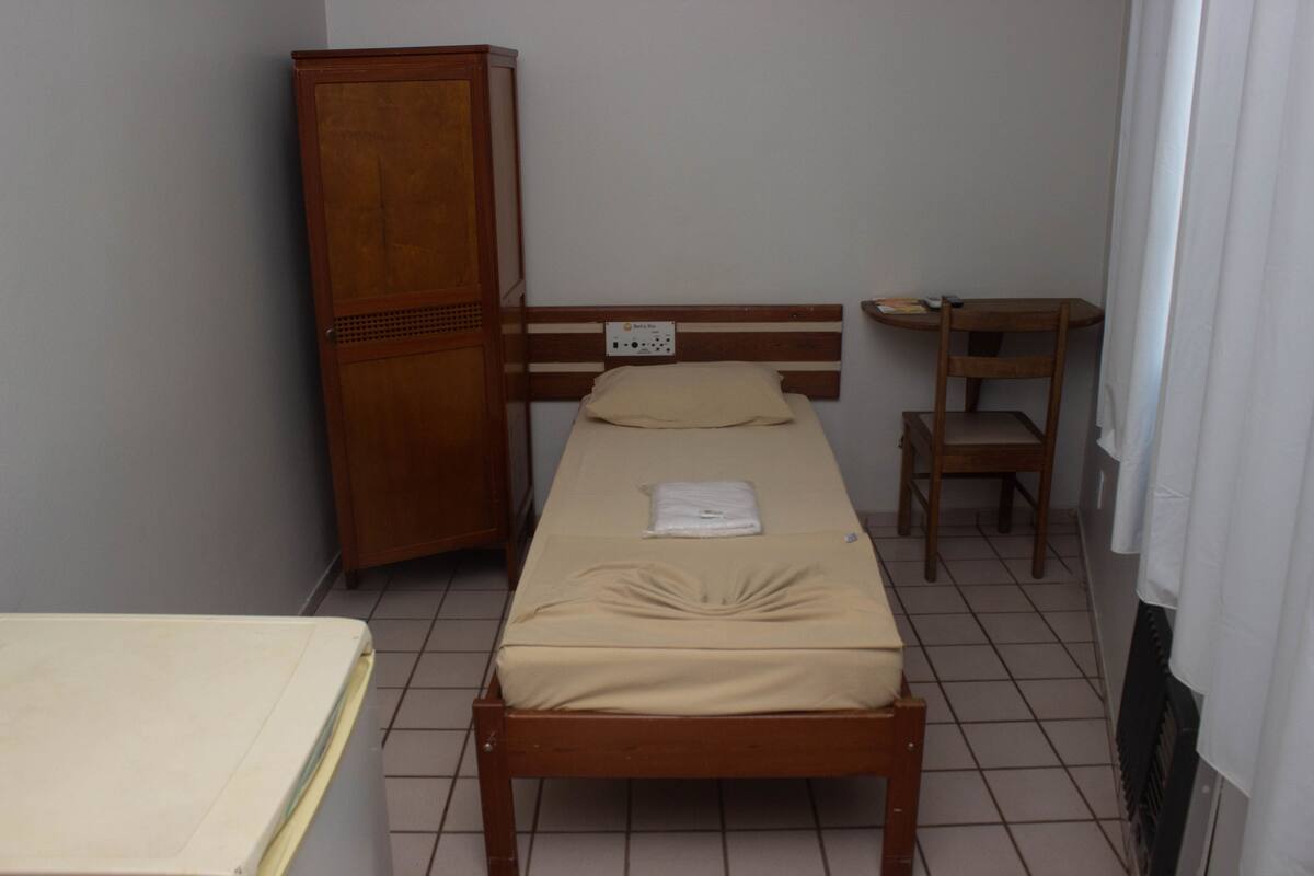 Ein kleines Zimmer mit einem Einzelbett, einem Holzkopfstück, einem Schrank aus Holz, einem kleinen Tisch und einem Stuhl.