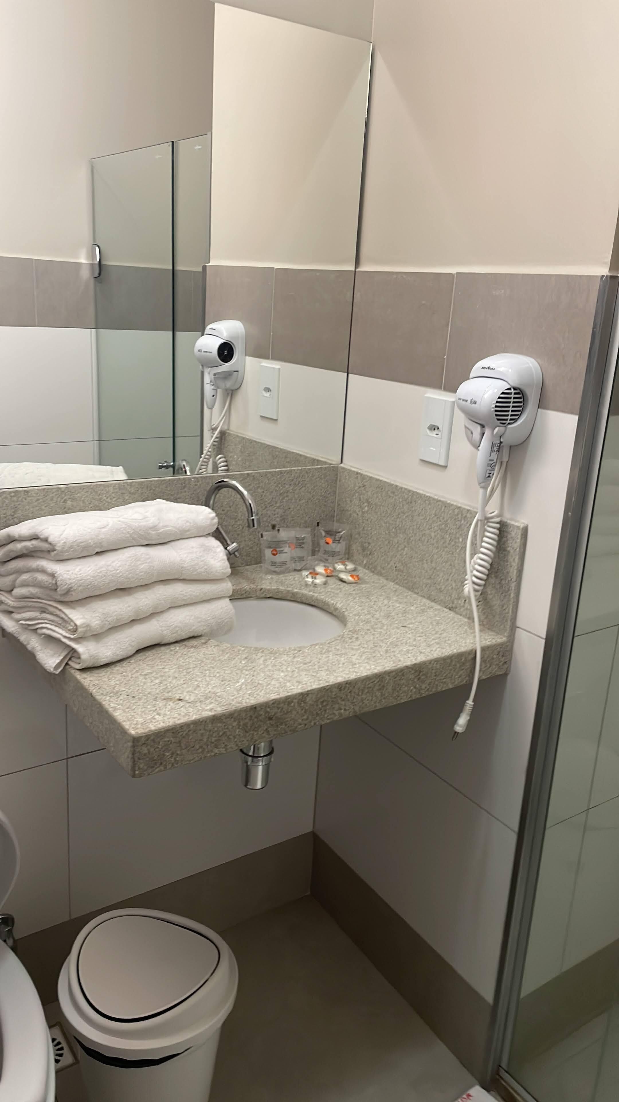 A bathroom with a granite sink, two hair dryers mounted on the wall, a stack of folded towels, and a trash bin.