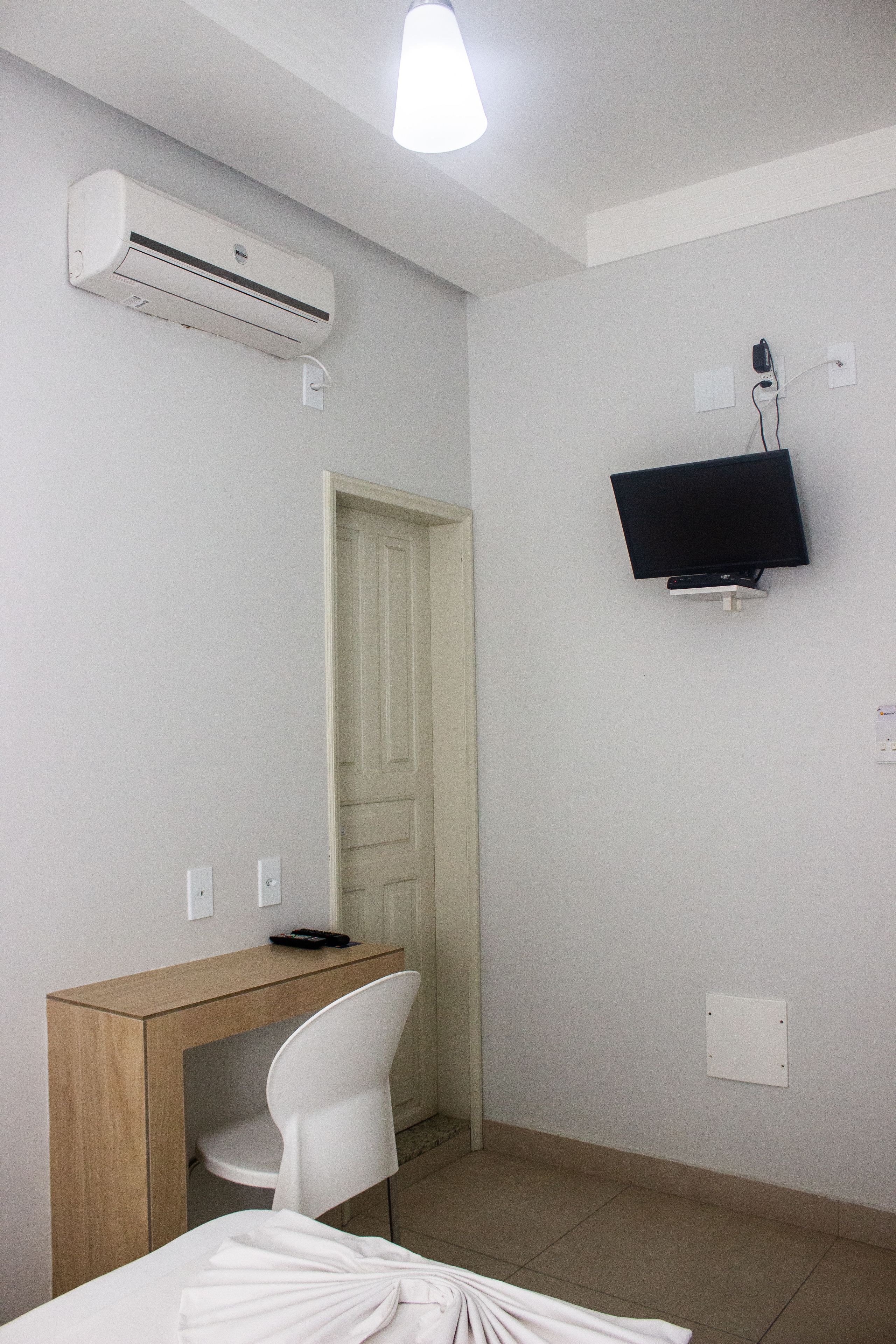 A hotel room with a desk, chair, bed, air conditioning unit, television, and a door.