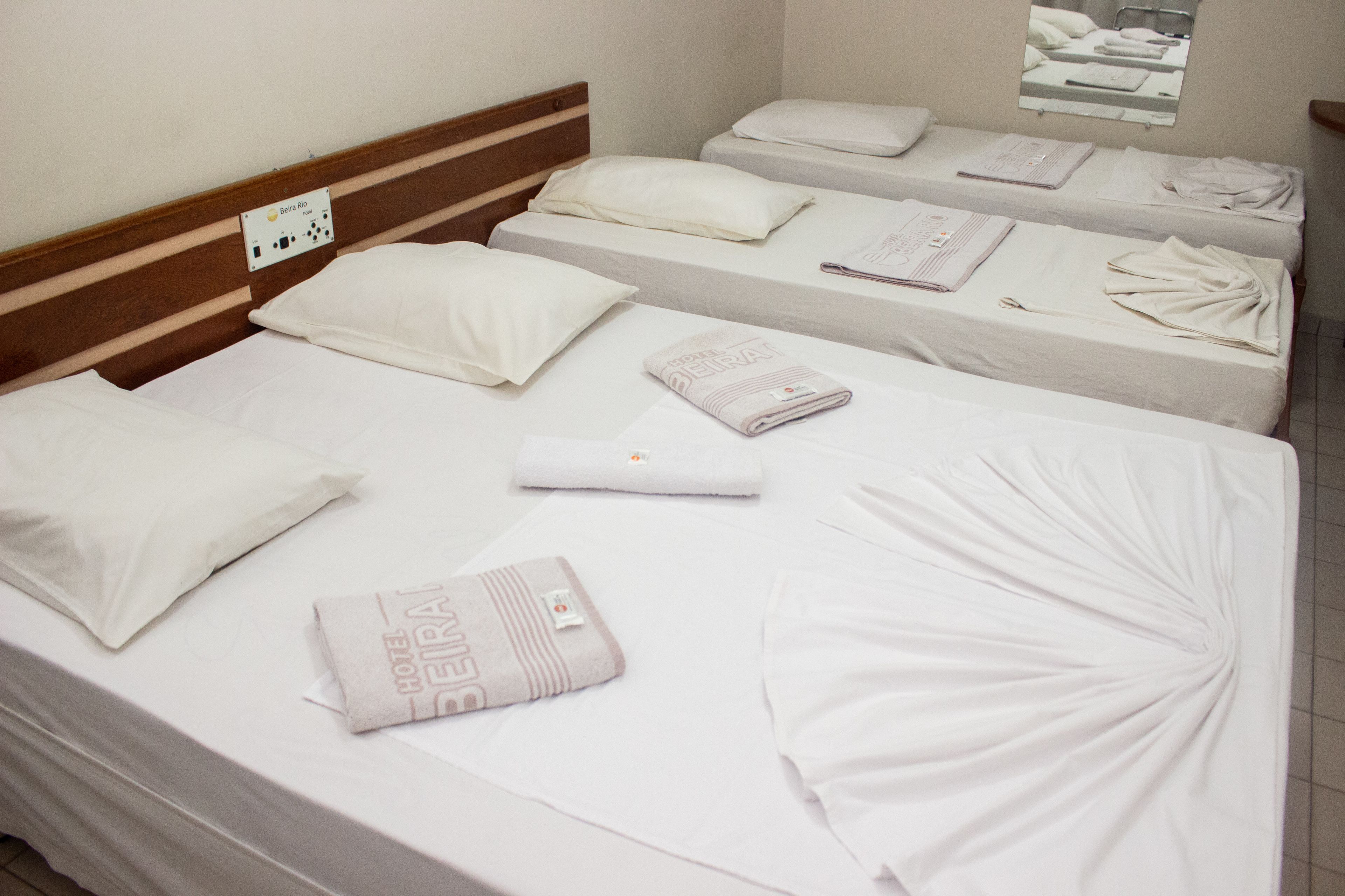 Quarto de hotel com três camas de solteiro, cada uma com colcha branca, almofadas e toalha dobrada. Possui cabeceira de madeira e uma pequena janela com vista para outro quarto.