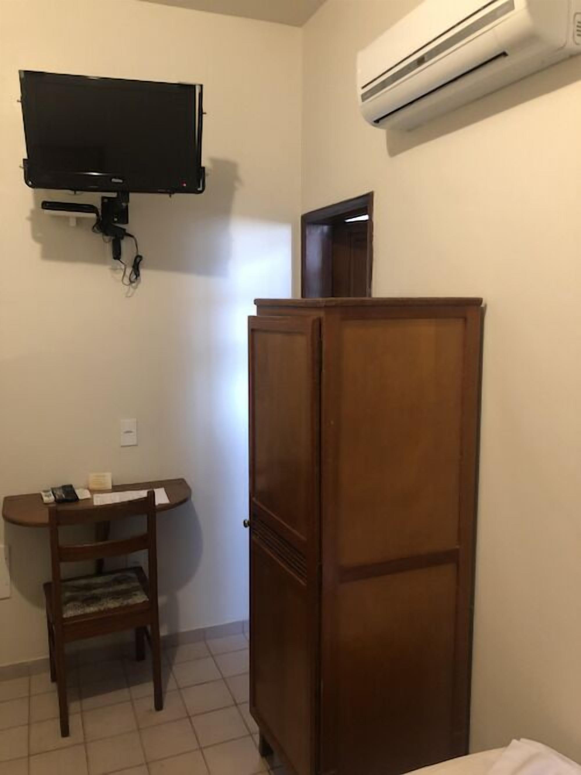 Une pièce avec un téléviseur fixé au mur, un meuble en bois, un bureau avec une chaise et un appareil de climatisation.