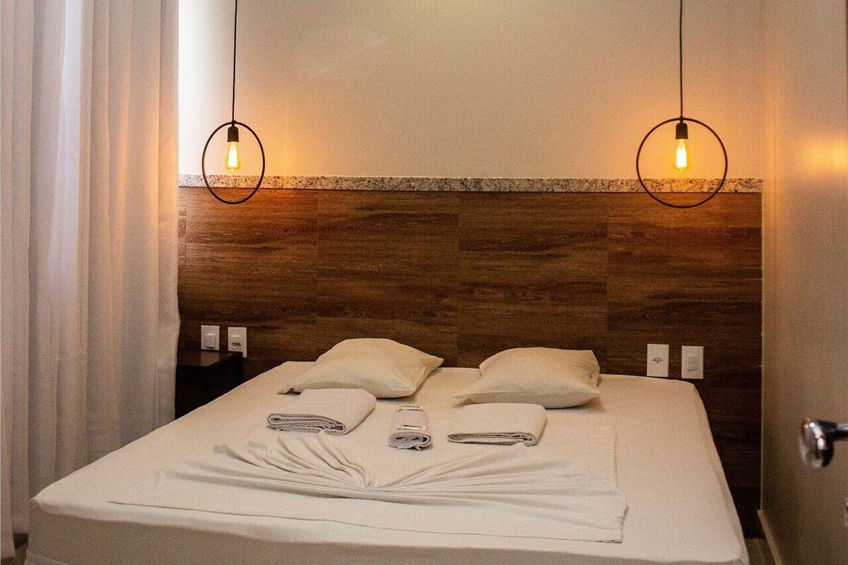 Ein Hotelzimmer mit einem Bett, zwei Pendelleuchten, einem Holzkopfteil und einem Fenster mit Vorhängen.