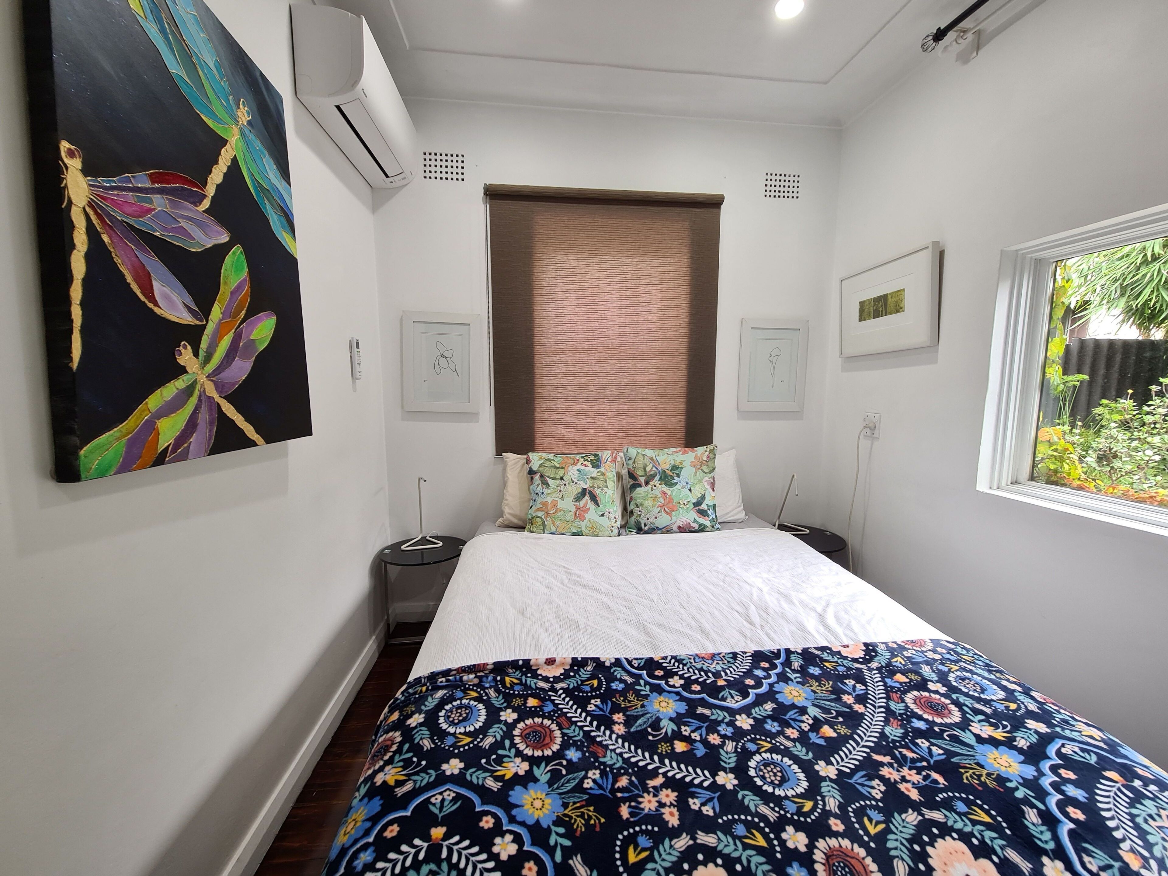A bedroom with a bed, a colorful artwork on the wall, an air conditioner, a window with a view of greenery, and a door with a handle.