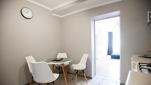 Un comedor con una mesa redonda y cuatro sillas blancas, un reloj en la pared y vistas a un balcón a través de una puerta abierta.