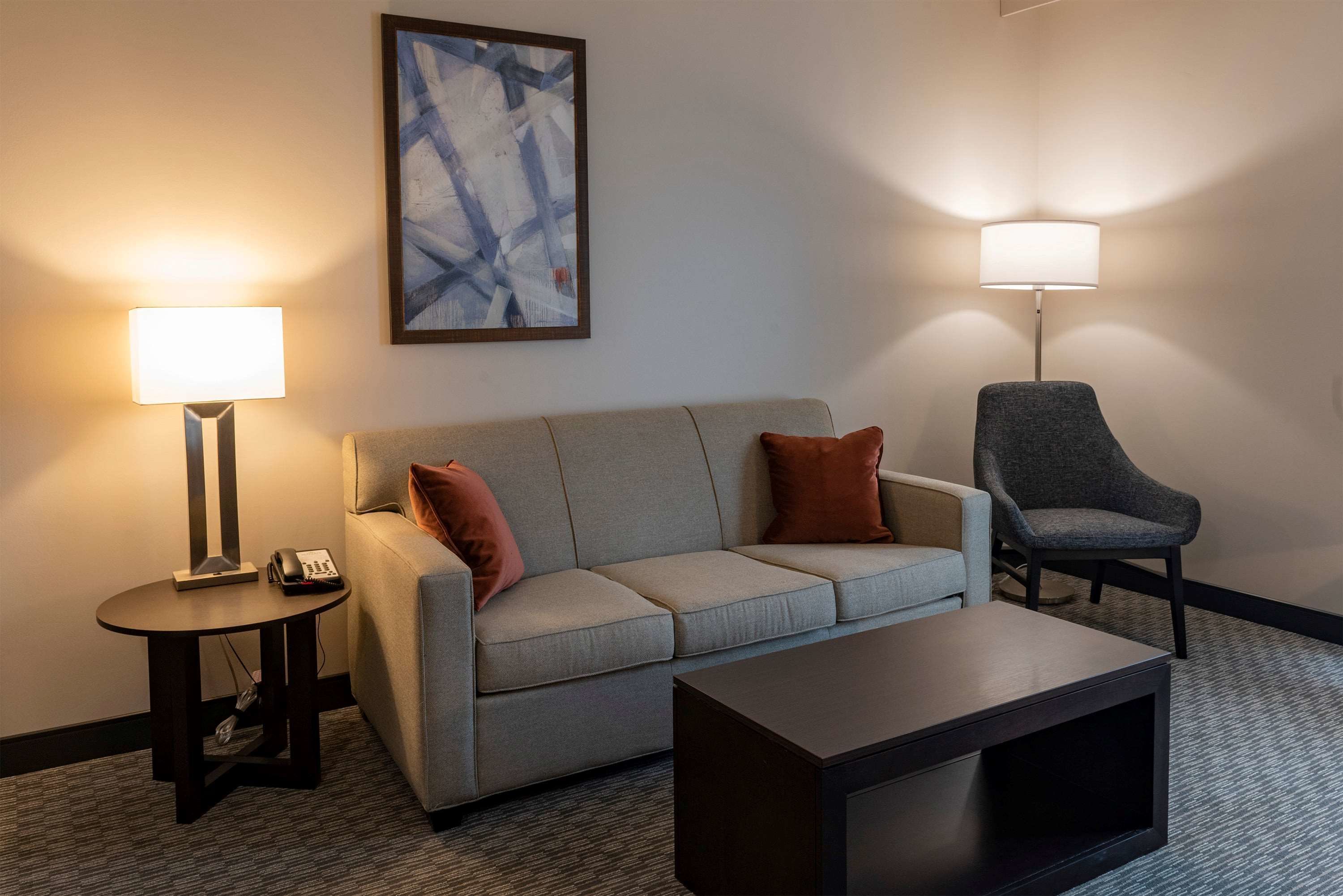 Une chambre d’hôtel moderne avec un canapé gris, une petite table ronde avec une lampe, un fauteuil sombre et un grand tableau abstrait accroché au mur.