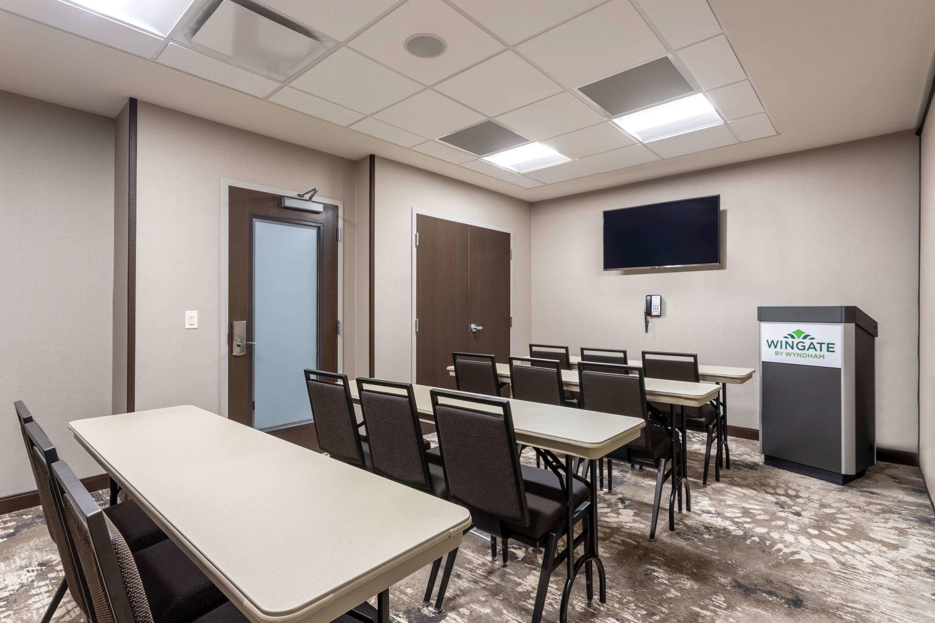 A conference room with multiple tables and chairs, a television mounted on the wall, and a podium with the logo "Wingate by Wyndham".