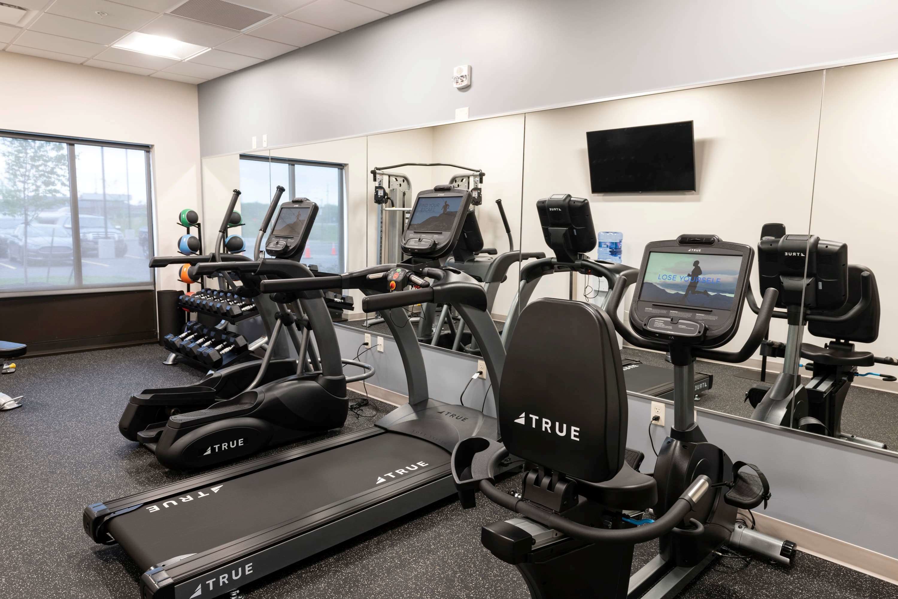 A gym with multiple exercise machines, including treadmills and elliptical trainers, and a large mirror reflecting the equipment.