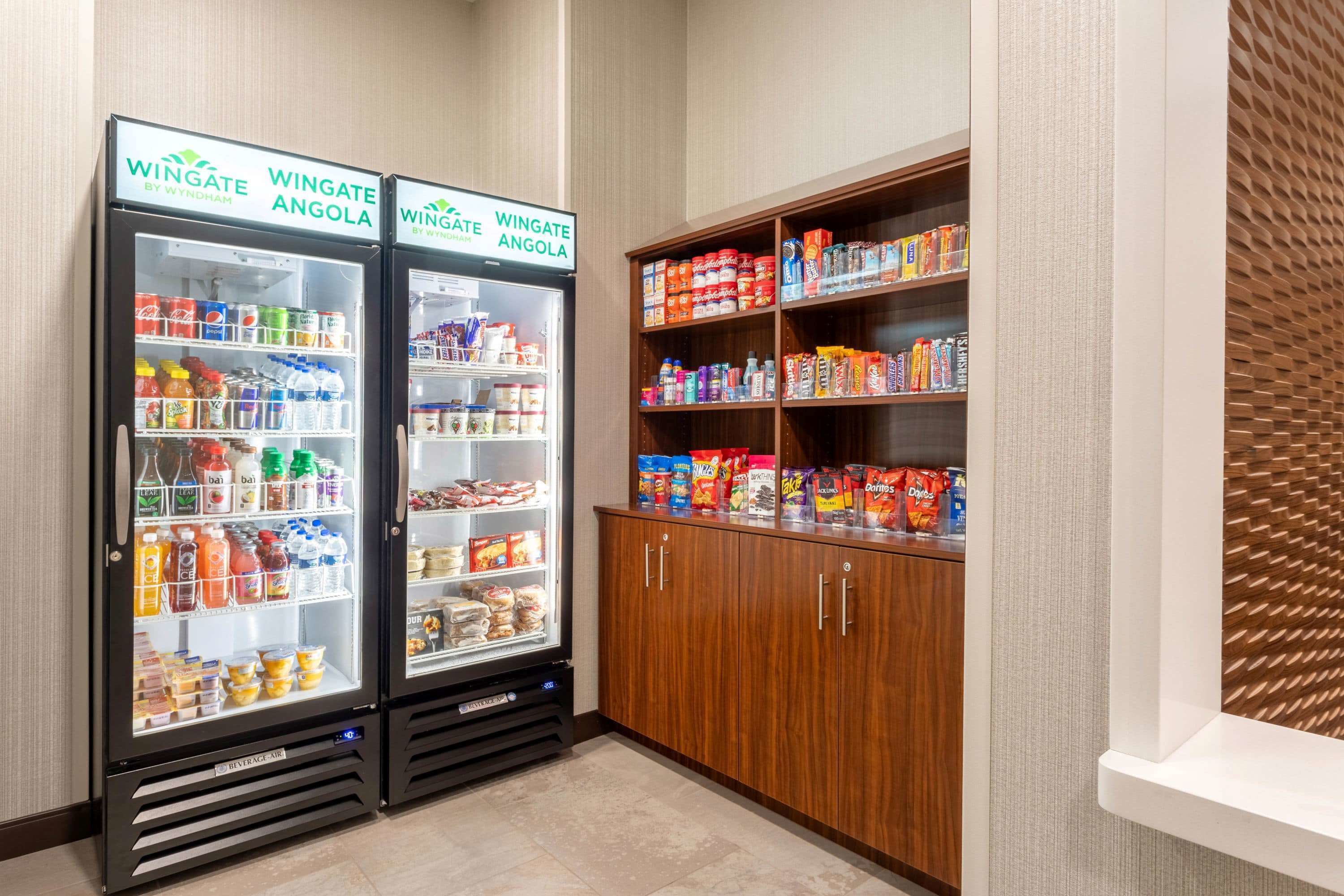 Two Wingate by Wyndham refrigerated beverage dispensers and a wooden pantry stocked with snacks and beverages.