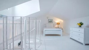 Un dormitorio moderno con decoración blanca, que incluye una cama, mesita de noche, lámpara y cómoda.