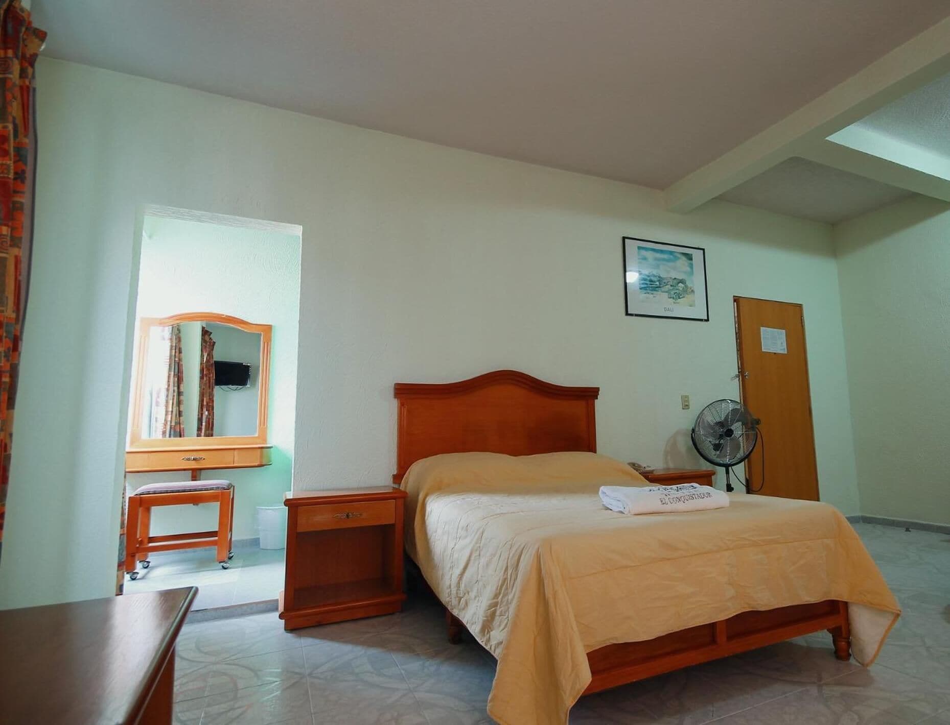 A bedroom with a bed, a wooden nightstand, a small table, a mirror, a fan, and a door.