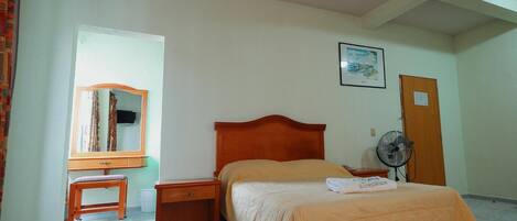 A bedroom with a bed, a wooden nightstand, a small table, a mirror, a fan, and a door.