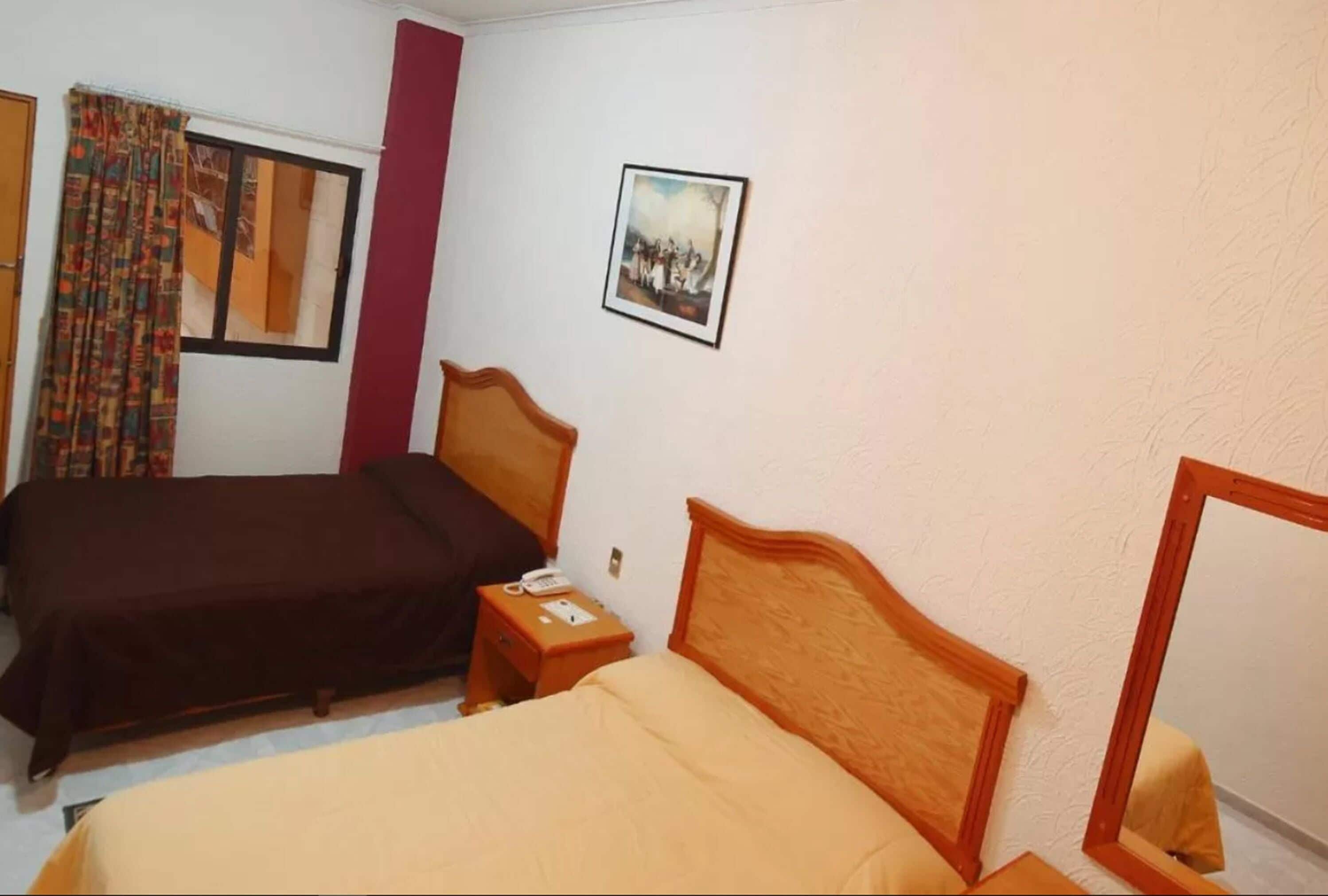 A hotel room with two beds, a window with curtains, a bedside table with a phone, and a framed picture on the wall.