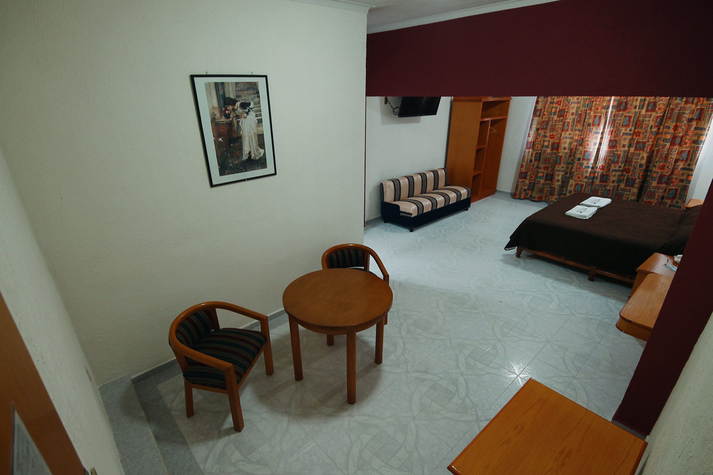 A hotel room with a bed, a sofa, a dining table with chairs, a TV mounted on the wall, and a patterned curtain.