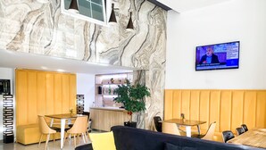 Un lobby de hotel moderno con paredes de mármol, un área de estar central con un sofá negro y cojines amarillos, y una zona de bar con estantes llenos de botellas.