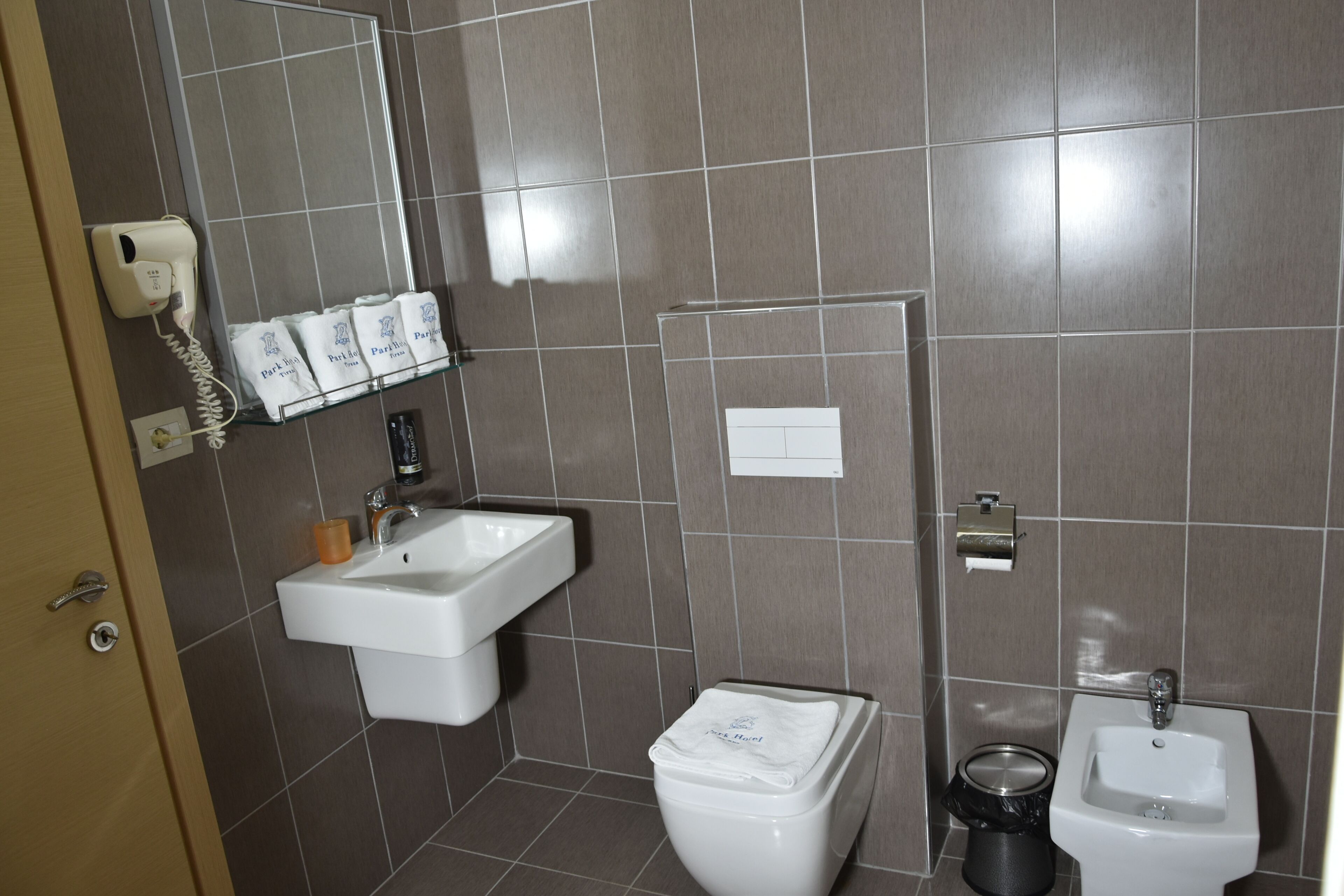 Une salle de bain avec un lavabo blanc, des toilettes et un bidet. Un sèche-cheveux est fixé au mur, et une étagère contient des articles de toilette.