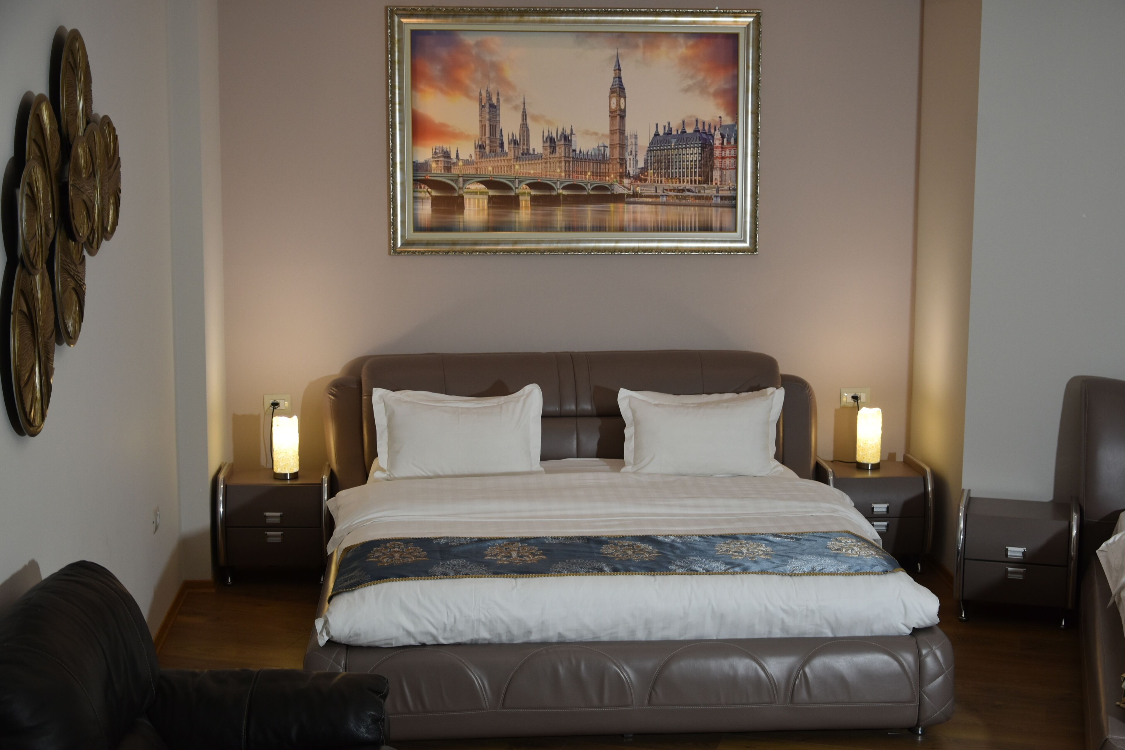 Ein Hotelzimmer mit einem großen Bett, zwei Nachttischen, einem Beistelltisch, einem Sessel und einem Stadtbild an der Wand.